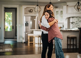 Pair of women embracing inside home with kitchen in background.