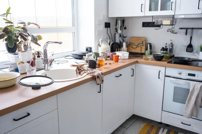 A messy kitchen counter with eggs, flour, bowls, oven mitts, cooking trays, and various utensils.