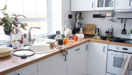A messy kitchen counter with eggs, flour, bowls, oven mitts, cooking trays, and various utensils.