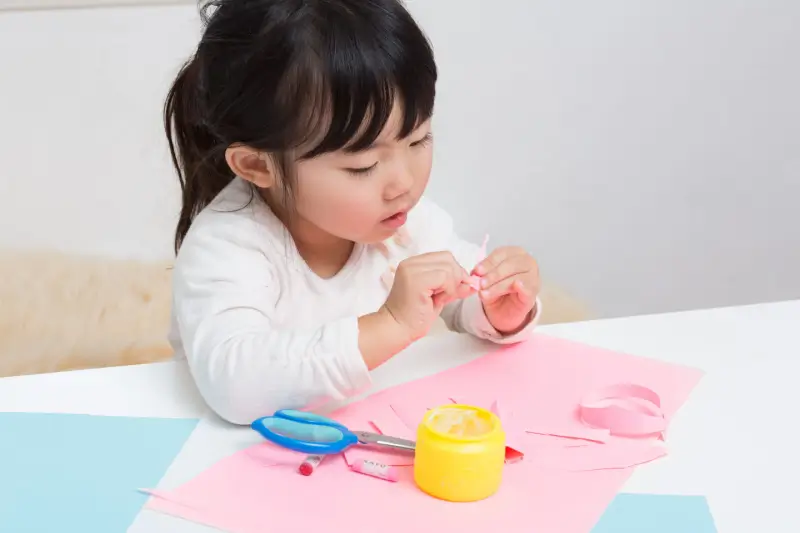 A young girl gluing pieces of pink paper.
